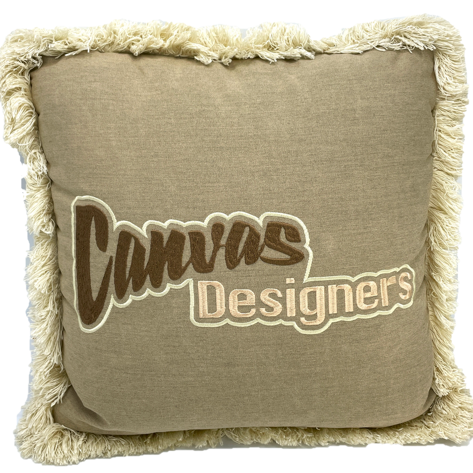 Fringed tan pillow embroidered with the Canvas Designers logo