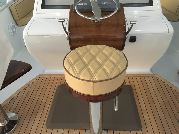 Round tan diamond-quilted helm stool on a pedestal in front of a wood helm and teak deck